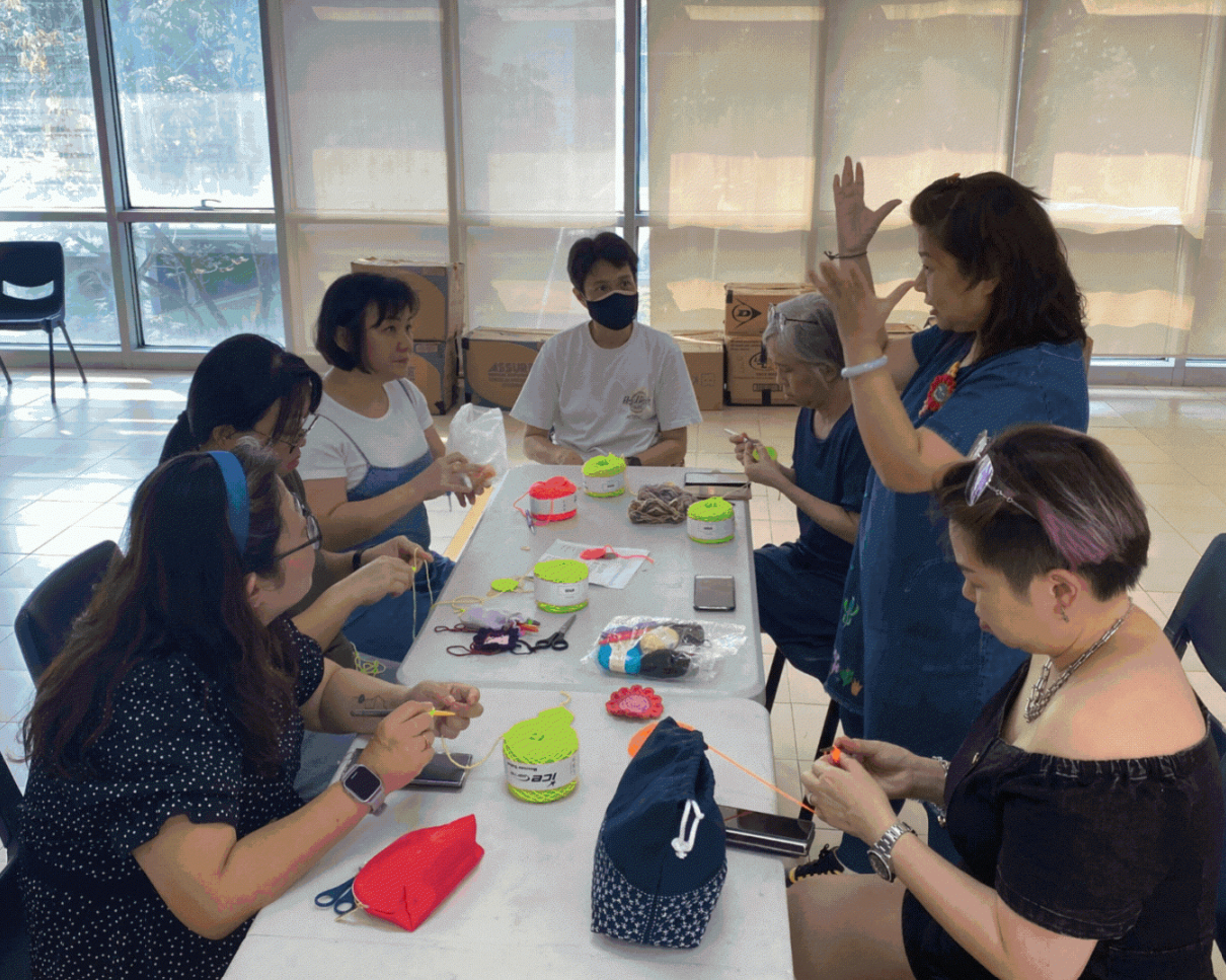 Group of people seated around a table doing crochet, with colourful yarn spools. One person stands, gesturing while explaining a technique.